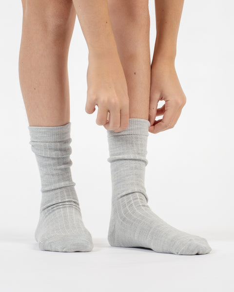 A front-facing view showing hands adjusting the top cuff of light gray ribbed merino crew socks on a person's legs.