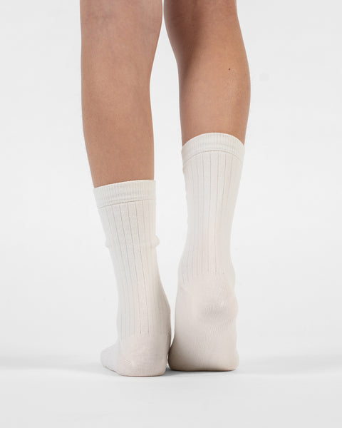 A back view of a person's lower legs and heels, standing flat in natural ribbed merino crew socks.
