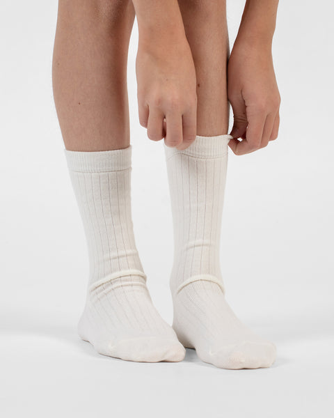 A front-facing view showing hands adjusting the top cuff of natural ribbed merino crew socks on a person's legs.