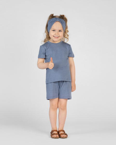 A young child smiles brightly while giving a thumbs-up, wearing a dusty blue short-sleeve t-shirt, matching shorts, and a knotted ribbon headband. They stand against a light gray background, completing the outfit with brown buckled sandals.