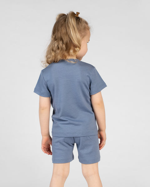A view from the back of a young girl with blonde hair in pigtails. She is wearing a dusty blue merino short-sleeve t-shirt and matching dusty blue merino shorts, standing against a plain light gray background.