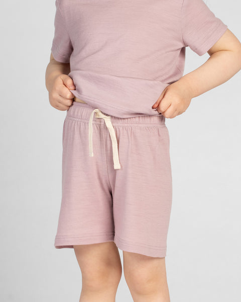 A close-up view of a young child's torso and upper legs, facing slightly to the side. The child is wearing a dusty pink merino short-sleeve t-shirt and matching dusty pink merino shorts. The child's hands are holding the hem of the t-shirt near the waist, revealing the elastic waistband and drawstring of the shorts.