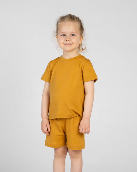 A young girl with blonde hair stands facing the camera, wearing a mustard yellow merino short-sleeve t-shirt and matching mustard yellow merino shorts. She is against a plain light gray background.