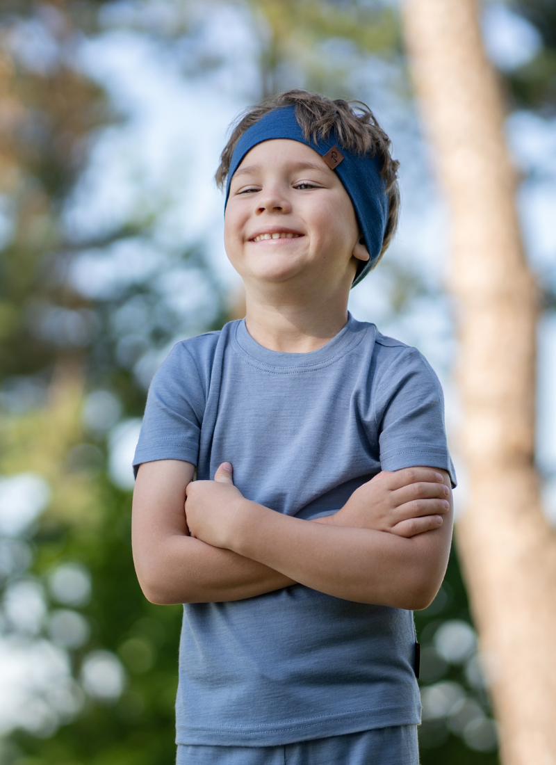 A smiling young boy standing outdoors with his arms crossed, wearing a dusty blue short-sleeved t-shirt and a matching blue headband, with blurred trees in the background.