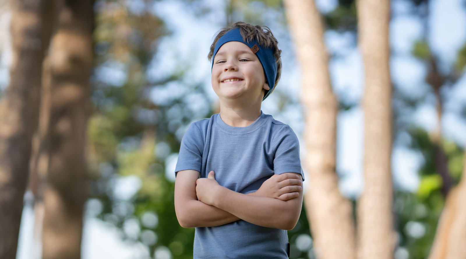 A smiling young boy standing outdoors with his arms crossed, wearing a dusty blue short-sleeved t-shirt and a matching blue headband, with blurred trees in the background.