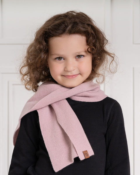A young child with curly brown hair and a slight smile looks directly at the viewer. They are wearing a black long-sleeved shirt and a light pink scarf with a small brown label, draped around their neck. The background is a white paneled wall.
