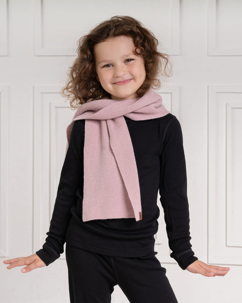 A young child with curly brown hair and a slight smile looks directly at the viewer. They are wearing a black long-sleeved shirt and a light pink scarf with a small brown label, draped around their neck. The background is a white paneled wall.