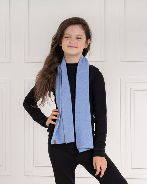 A young child with long dark brown hair, stands with one hand on their hip and a slight smile. They are wearing a black long-sleeved shirt, black pants, and a light blue knit scarf draped around their neck. The background is a white paneled wall.