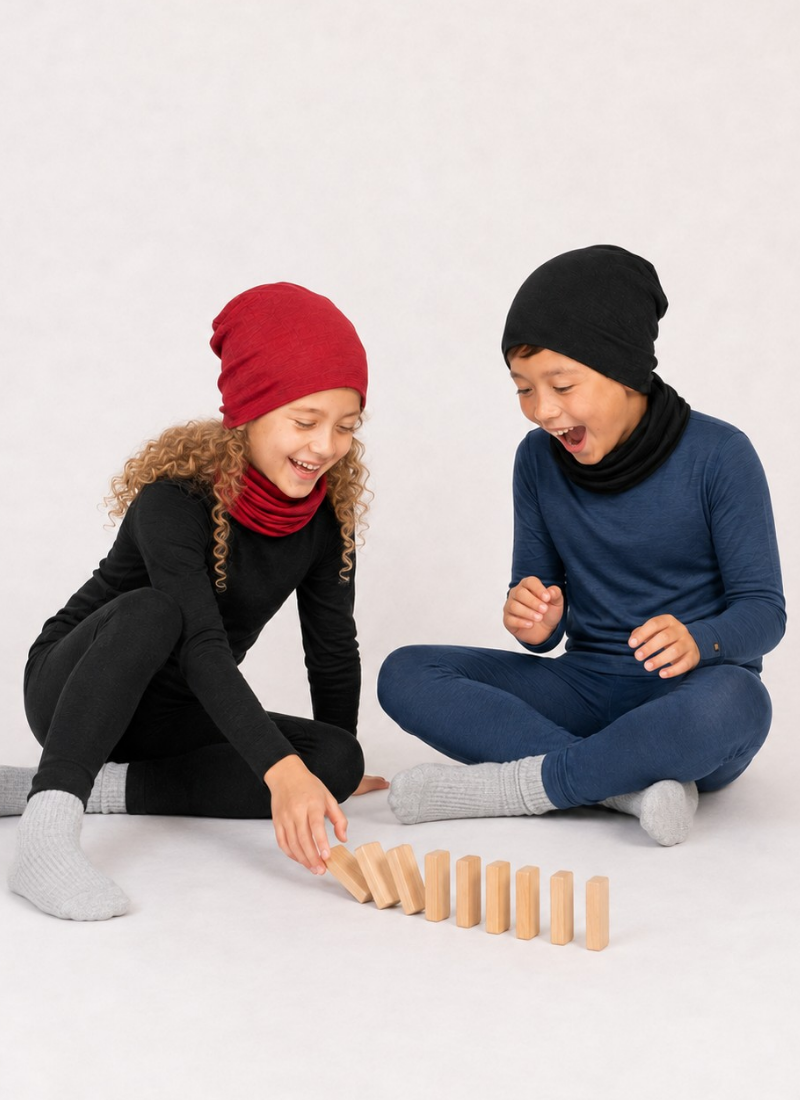Two children smiling and sitting on a white floor playing with wooden dominoes. The girl on the left is dressed in a black long-sleeved base layer set, a dark red beanie, a matching red neck gaiter, and grey ribbed socks. The boy on the right wears a navy blue long-sleeved base layer set, a black beanie, a matching black neck gaiter, and grey ribbed socks.