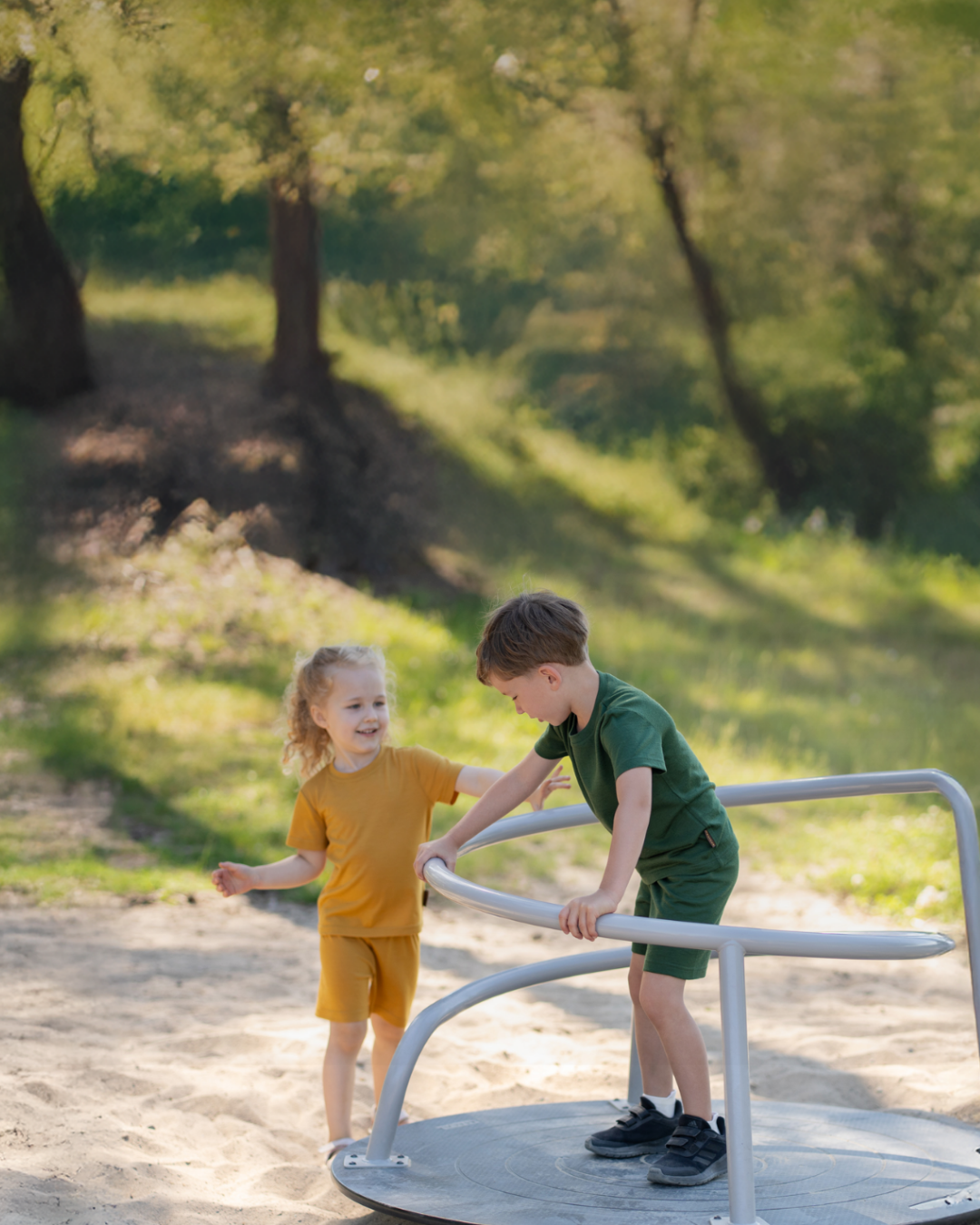 Two young children—a girl in a yellow outfit and a boy in a dark green outfit—playing together on a metal spinning merry-go-round in a sandy playground surrounded by trees.