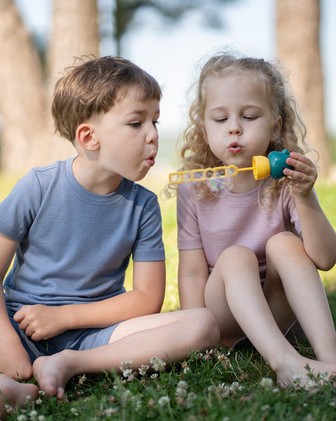 Two children sitting together on the grass wearing Merino wool T-shirts and shorts while blowing bubbles on a sunny day. The lightweight, breathable kids' outfits are showcased in a playful outdoor setting, perfect for everyday comfort and active adventures.