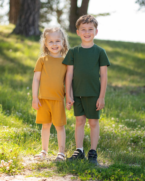 Two children standing hand in hand outdoors wearing matching Merino wool T-shirts and shorts in yellow and dark green. The lightweight, breathable kids' outfits are showcased in a sunny park setting, perfect for everyday play, family outings, and warm-weather adventures.