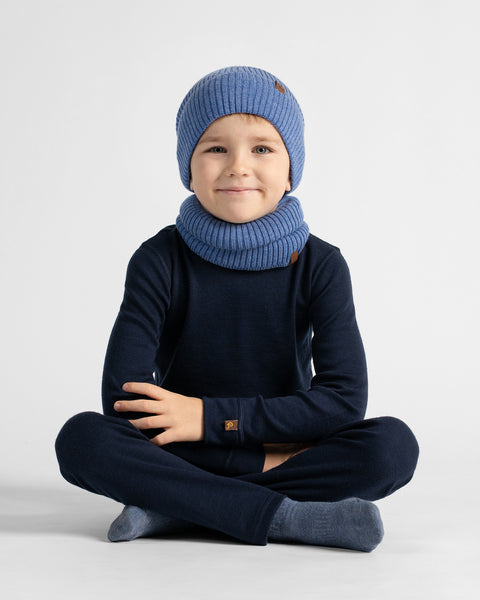 A child sitting cross-legged on the floor against a plain background, wearing a dark navy long-sleeve base layer set with a bright blue beanie and matching neck warmer.