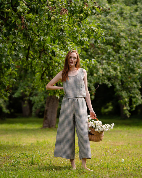 The model in the photograph is wearing mid-rise, light green and white gingham pattern wide leg pants. The culottes are matched with a sleeveless top in the same color option. She is holding a basket full of flowers, standing under a tree barefoot on the grass.