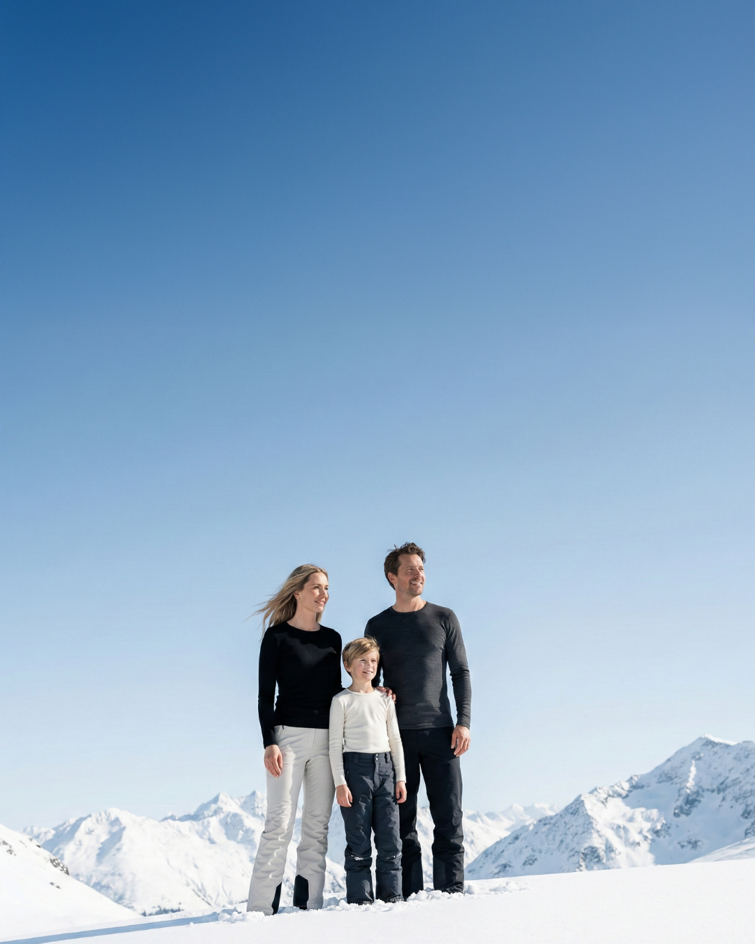 Family of three standing on a snowy mountain peak, wearing black base layer tops and thermal pants, with alpine mountains in the background.