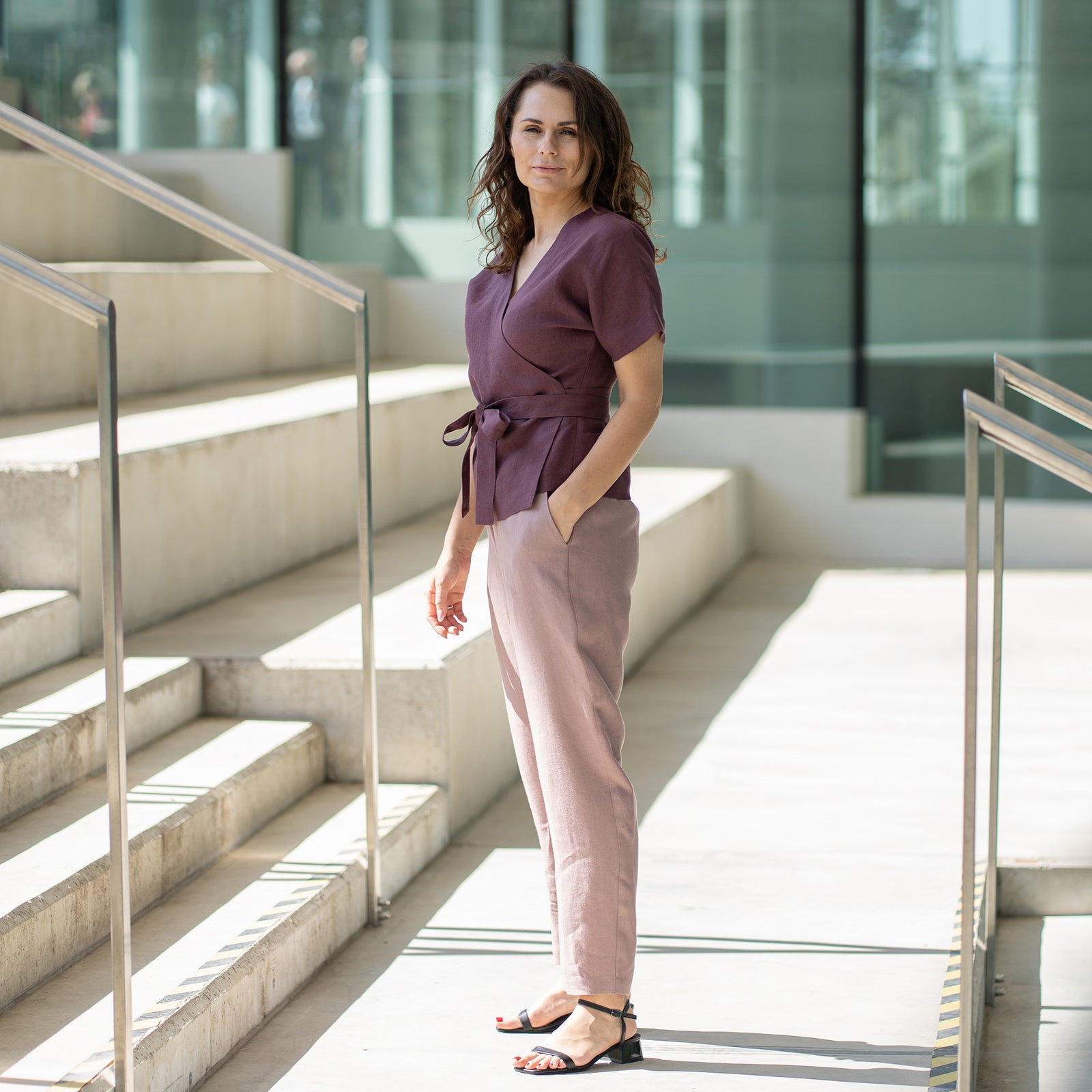 A woman with brown hair, wearing a dark purple short-sleeved wrap top and faded rose trousers, poses on indoor concrete stairs.