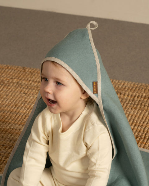 A close-up of a happy baby sitting on a woven rug, looking off to the side with an open mouth. The baby is wearing a light cream-colored top and a dusty sage green hooded towel with light beige binding. A small brown logo tag is visible on the towel's edge. The background is a solid, warm taupe wall.