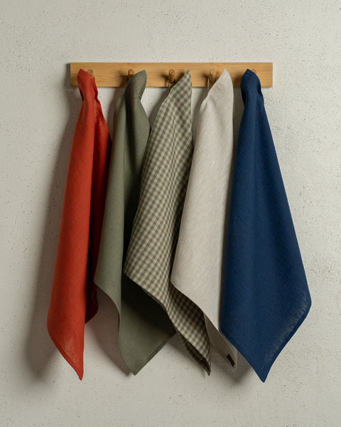 The menique square linen dish towels in the colors cinnamon red, stone green, green check, natural, storm blue hanging on a wooden rack on a wall.