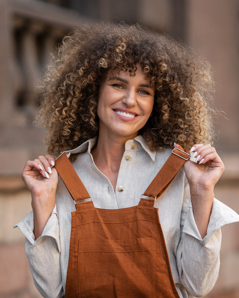 Woman with curly hair standing in a street and wearing Linen Pinafore Jumpsuit Nicci Almond Brown