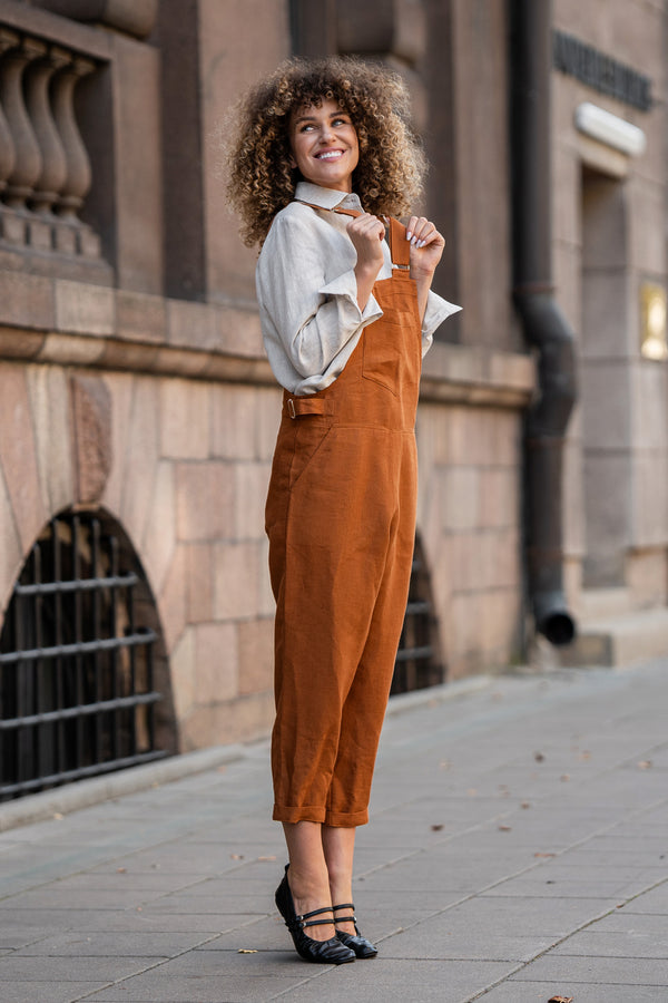 Woman with curly hair standing in a street and wearing Linen Pinafore Jumpsuit Nicci Almond Brown