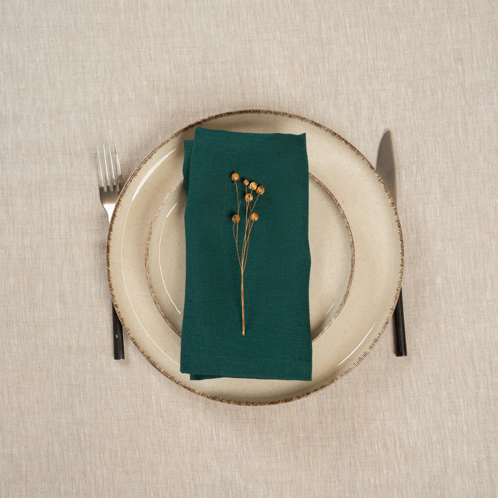 A tabletop covered with a linen tablecloth, with a vase and a single plate set on top of it. By the sides of the plates there are cutlery items, and there is a linen napkin in cobalt blue color on top.