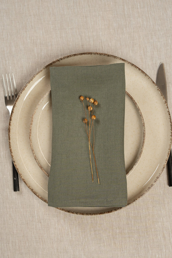 The menique linen napkin in stone green color, put on top of two stacked plates . There are cutlery items by the sides of the plates. The background is a natural color linen tablecloth.