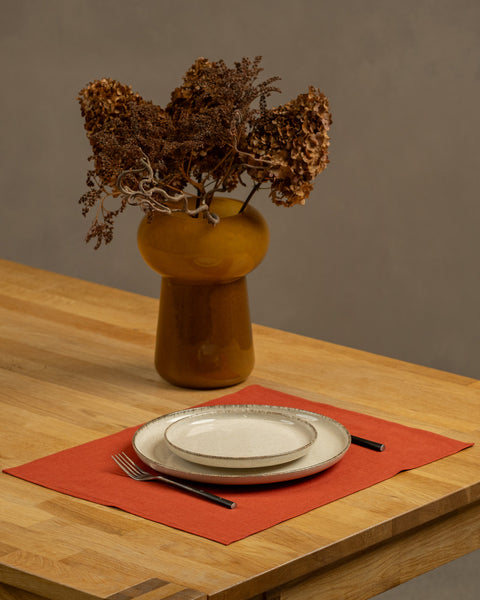 The menique cinnamon red linen placemat on a light wooden table. There are two different size porcelain plates and cutlery on top of it. There is also a vase on the table.