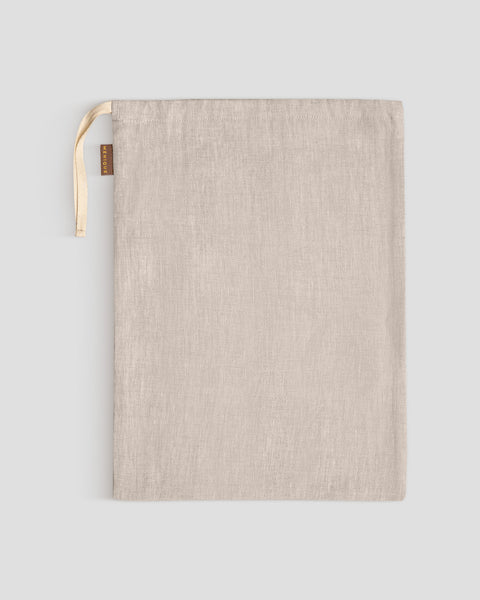 A menique linen storage bag lies flat on a light gray surface, its light-colored drawstrings gathered at the top left, next to a small brown tag.