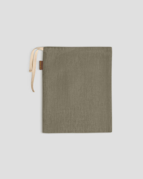 A stone green linen drawstring bag lies flat on a light gray surface, its light-colored drawstrings gathered at the top left, next to a small brown tag.