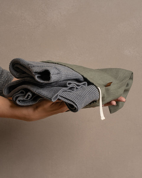 A person&#39;s hands gently hold a folded gray knitted garment inside a stone green linen drawstring bag against a muted brown background.