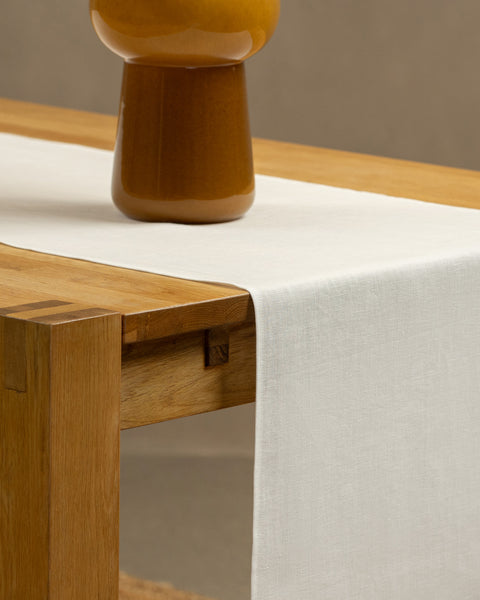 The pure white linen table runner on top of a wooden table, captured in a close up picture from the corner of the table. 