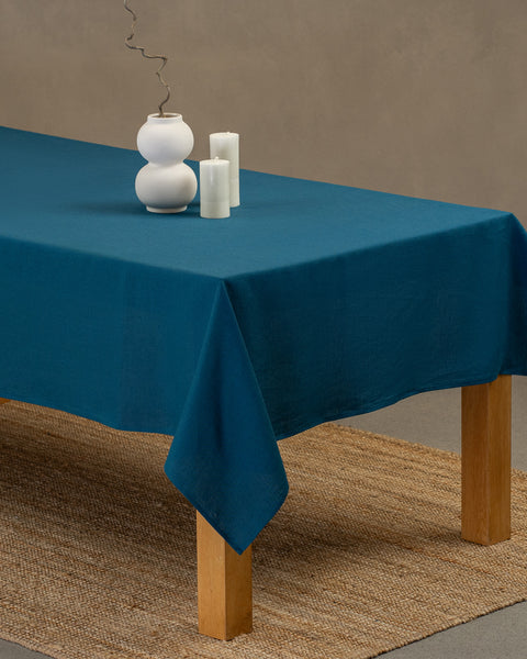 The menique linen tablecloth in cobalt blue color, draping over a wooden table, with a modern white vase on top of it and two different height white candles by the sides.