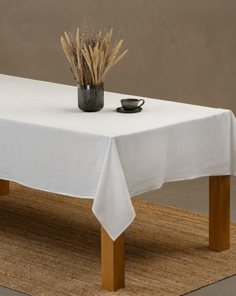 The menique linen tablecloth in pure white color, draping over a wooden table, with a glass vase and a teacup with a plate on top of it.