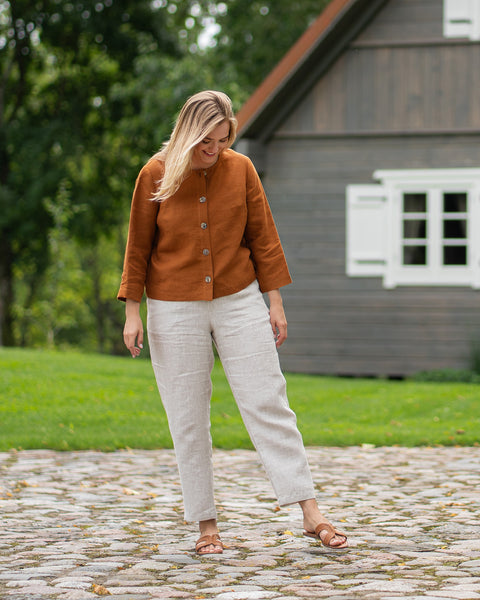 A full-length portrait shows a woman standing on a gravel path outside a wooden house. She is wearing an orange top and white pants while looking slightly downwards.