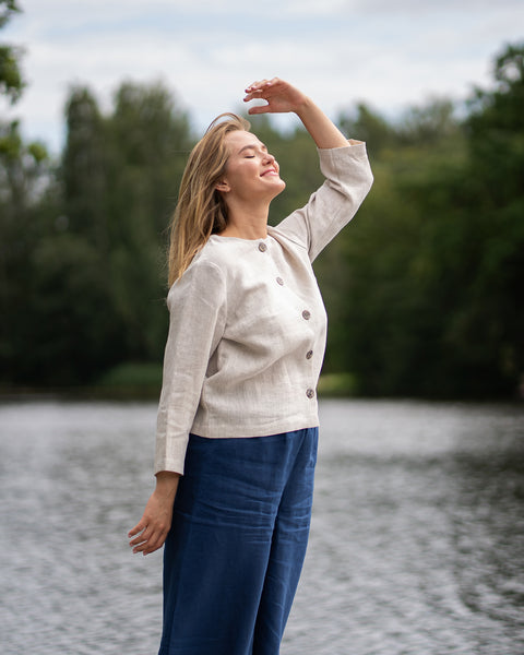 A blonde woman standing by the water tilts her head upwards and rests one hand lightly near her forehead. She is wearing a light sweater and blue pants against a blurred background of green trees.