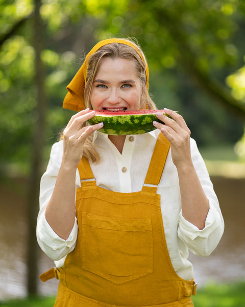A smiling woman wearing a mustard yellow overall dress and matching headwrap stands outdoors in a lush, green park. She is cheerfully holding up a slice of fresh watermelon to take a bite.