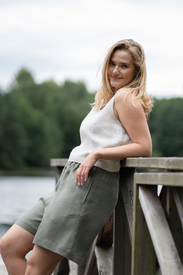 A smiling woman leans comfortably against a wooden pier railing, looking directly at the camera with her hair gently blowing in the wind. She is wearing a natural linen sleeveless top and stone green bermuda shorts, set against a softly blurred background of a lake and green trees.