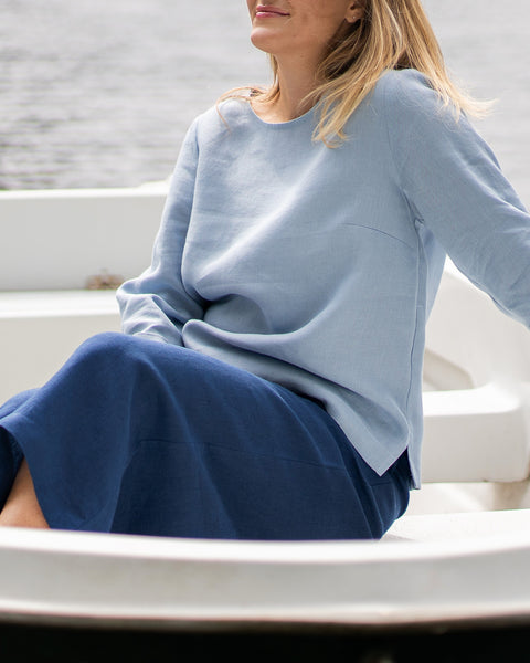 A woman sits casually in a small white boat, wearing a light blue, long-sleeved linen top and dark blue pants. Sunflowers are partially visible resting near her lap.