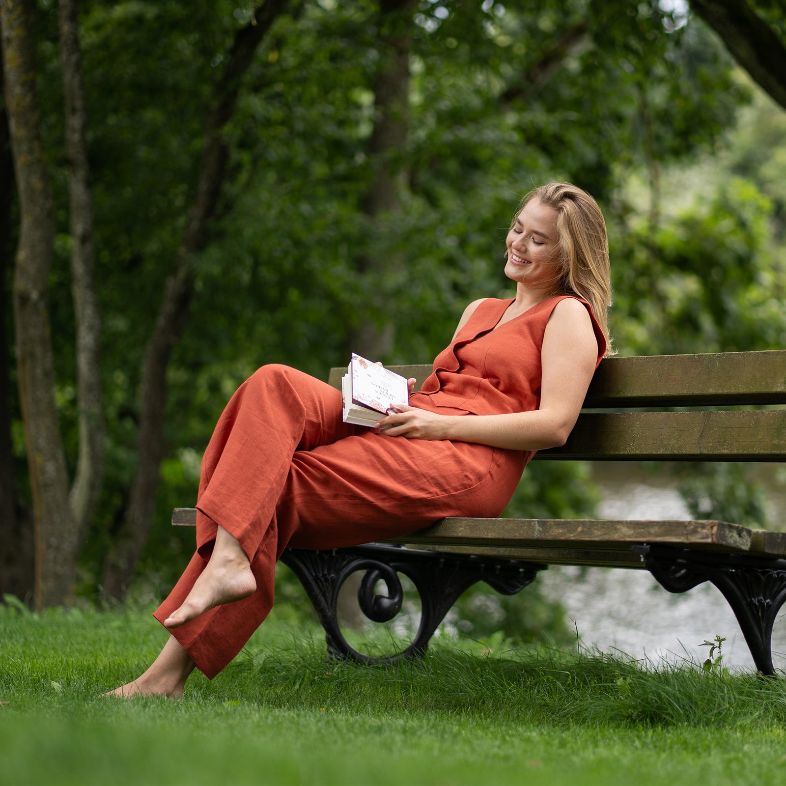 A fair-skinned woman models a cinnamon red colored linen vest and wide-leg trousers, standing against a textured light beige wall. She is wearing black sandals.