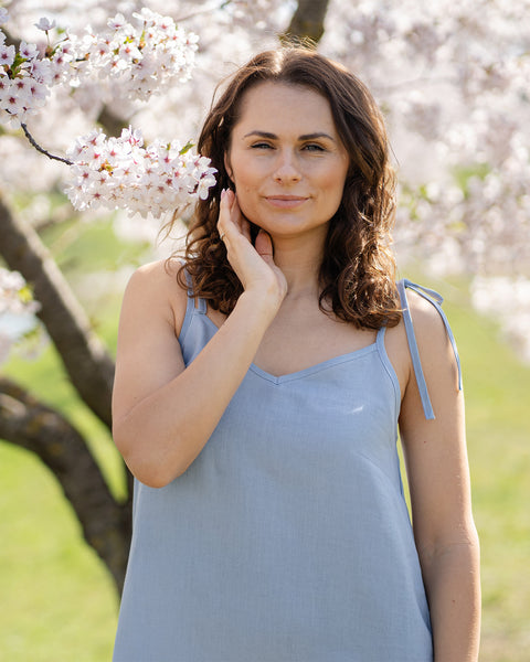 A woman wearing a cloudy blue linen slip dress stands beside a tree covered in white blossoms.