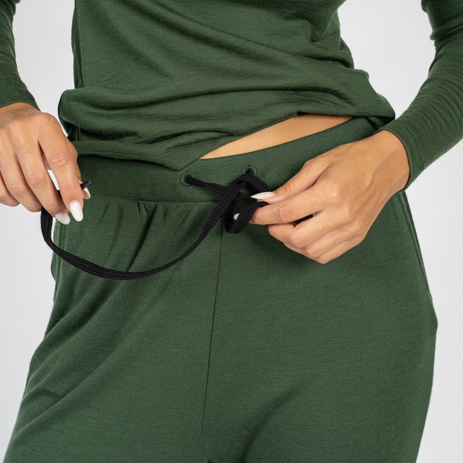 A young woman wearing the menique women's merino wool 250gsm jogger bottoms and long sleeve shirt set. Her hands is behind her head and another one by the hip, pulling up the shirt.