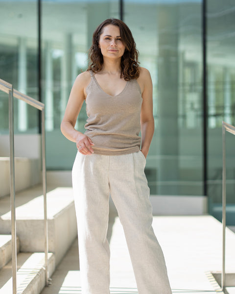 A woman with shoulder-length wavy brown hair stands looking directly at the camera, with one hand in her pocket. She wears menique tank top and wide-legged white pants Lotus, positioned on light-colored stairs with silver handrails. A modern building is in the background.