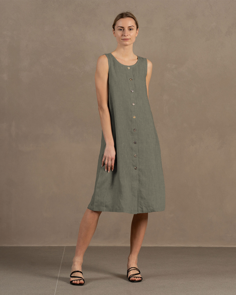 A light-skinned woman with her hair pulled back stands facing forward, modeling a sleeveless, stone green linen dress with a button-down front.