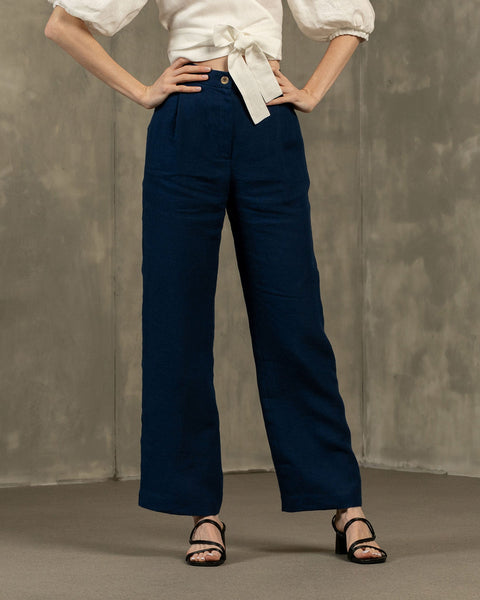 A person is shown from the waist down, modeling menique, high-waisted, wide-leg linen pants. They have their hands on their hips and are wearing black open-toe heeled sandals. The background is a textured gray wall and floor.