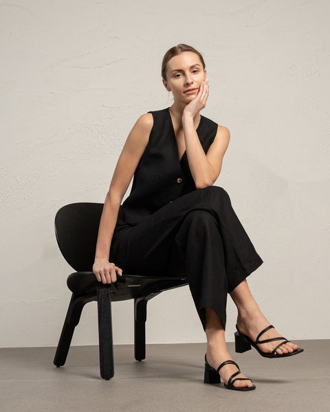 A woman with light skin and blonde hair pulled back sits thoughtfully on a black chair against a textured light gray wall. She rests her right elbow on her right knee and her chin on her right hand, gazing slightly to the left of the frame. She is wearing a menique linen vest, along with pure black wide-legged linen pants.
