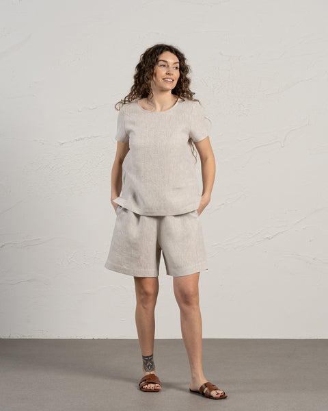 A woman with brown curly hair wearing the menique 100% linen bermuda shorts and linen t-shirt top set. Her hands are in the pockets, while she is looking sideways. The background of the photo is bright and in cool tones.