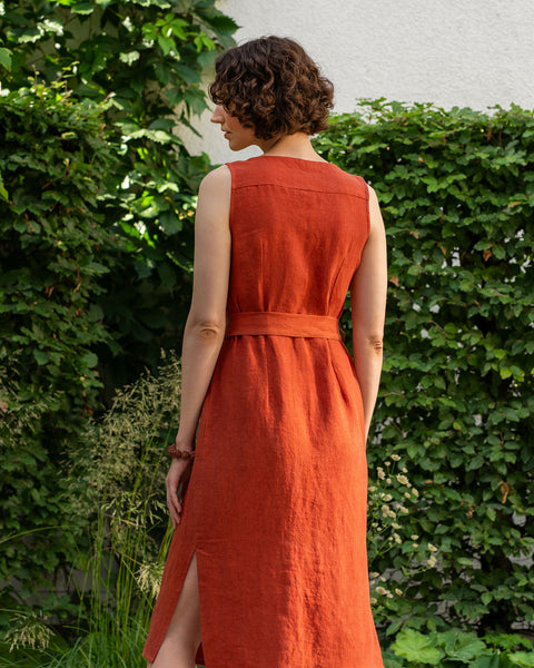 A fair-skinned woman with short, curly dark brown hair stands with her back mostly to the viewer, angled slightly to her left, outdoors. She is wearing a cinnamon red linen dress that reaches below her knees and has a slit on the left side. The dress is sleeveless, and a belt of the same fabric is tied around her waist. 