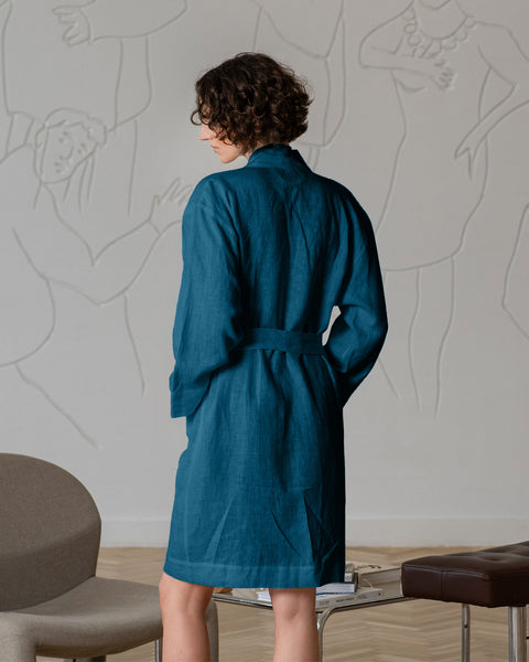 A woman with short, curly dark hair and glasses, wearing a menique linen robe tied at the waist, stands barefoot with their back to the viewer. A gray chair is to their left, and a table with a brown stool is to their right against a light gray wall with faint figure drawings.