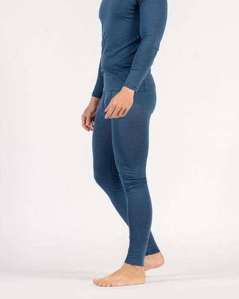 A side profile view of a person from the neck down standing barefoot against a white background, wearing a fitted, dark blue long-sleeved thermal top and matching thermal leggings. Their right heel is slightly lifted in a relaxed stance.