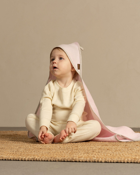 A baby sitting on a woven natural fiber rug, looking thoughtfully upwards. The baby is wearing a light cream-colored long-sleeved top and pants, and is draped in a soft, light pink hooded towel with grey binding. The background is a solid, warm taupe wall.